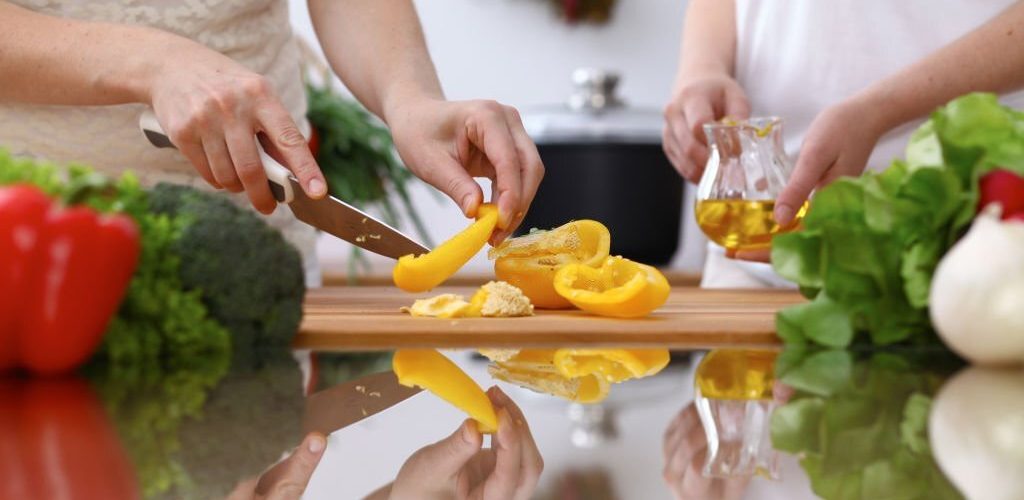 Closeup of human hands cooking in kitchen. Mother and daughter or two female cutting bell pepper for fresh salad. Healthy meal, vegetarian food and lifestyle concepts.
