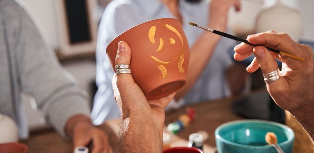 Cropped view of the senior man holding clay pot and brush while drawing patterns on it at the loft studio during the master class. Stock photo