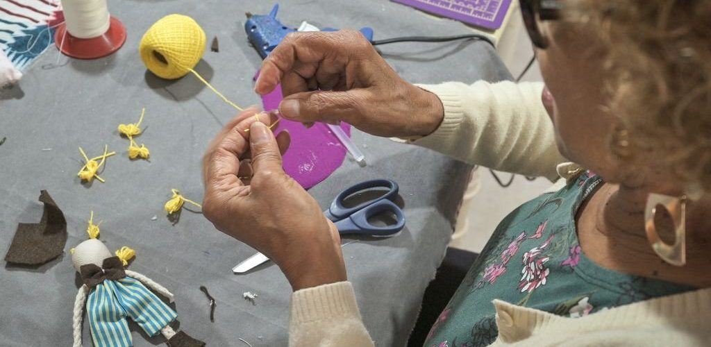 Portrait of an artisan woman at home in her atelier, handcrafting a cloth doll with sustainable materials and cloth remains. Around her there are some tools such as a pen, scissors and sewing threads, used for manufacturing the cloth doll.