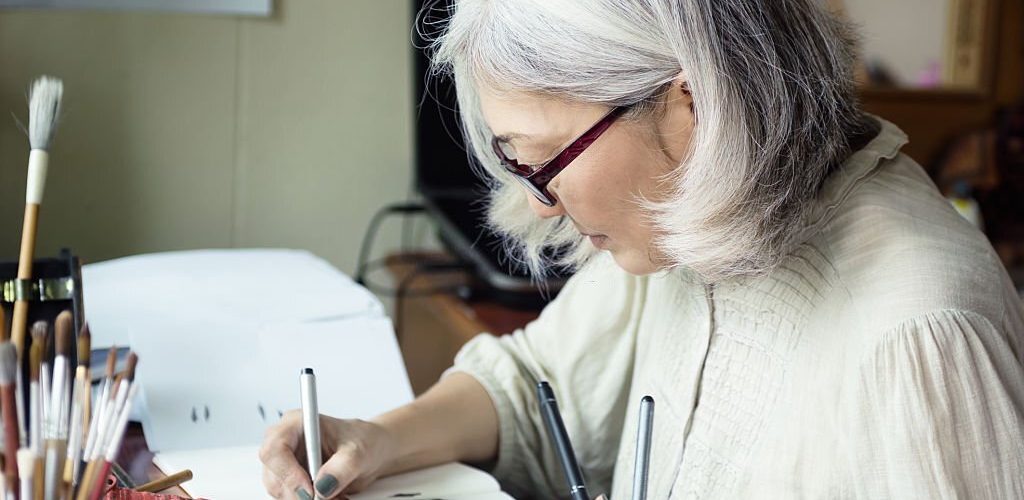 Asian senior woman artist sketching. Focus on face, blurred workspace in background. Art background, text space